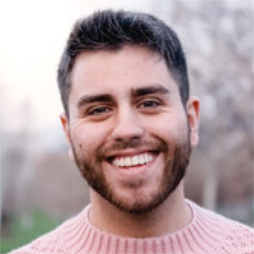Young man with dark hair and beard smiling at camera outdoors, wearing a pink sweater.